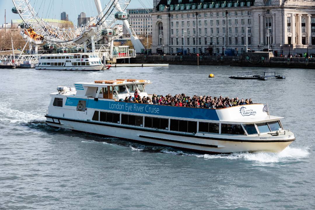 A Thames River Cruise boat with many people on top of it.