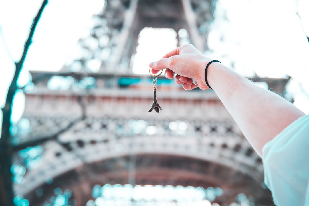 A person holding up a keychain of the Eiffel Tower in front of the Eiffel Tower.